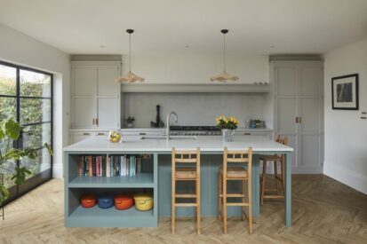 A classic shaker-style kitchen featuring a green and light grey cabinetry with brass hardware. The large island, painted in green, includes a bookshelf filled with cookbooks and colourful cast iron pots, topped with a white quartz worktop. Wooden bar stools provide seating at the island. The cooking area showcases a large range cooker with a large chimney and mantle above. Two pendant lights hang above the island, adding warmth and texture. Herringbone wood flooring and black-framed glass doors allow natural light to enhance the inviting space.