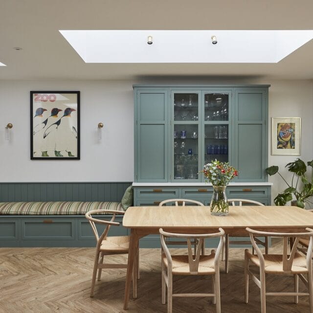 A classic shaker-style kitchen with a banquette seat with striped upholstery and a tongue and groove paneled back, with artwork and wall lights hanging above. The green-painted cabinets store glassware and other kitchenware behind glazed doors and wide drawers below. An oak dining table has Wishbone-style chairs. Plants and flowers create a an inviting space.
