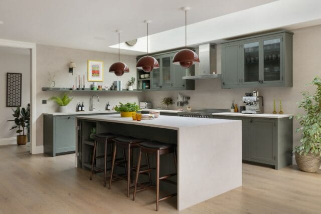 Shaker kitchen island with micro-concrete worktops and waterfall ends, illuminated by vermillion red flowerpot pendant lights. Features open shelving for cookbooks and ornaments, along with a slim wine cooler.
