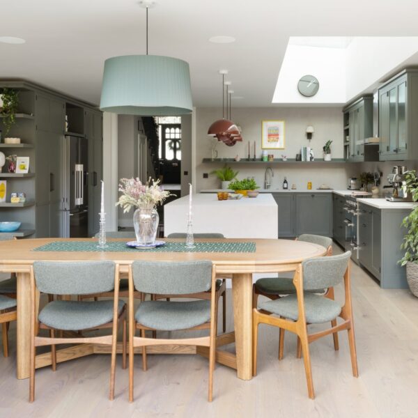 Large oval wooden dining table with a green pendant light above. Shaker kitchen behind with a large island with micro-concrete waterfall ends. Skylight floods the room with light.