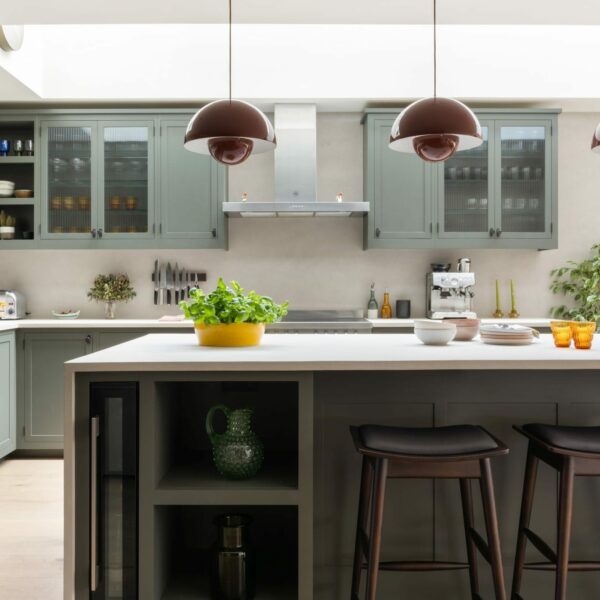 Shaker kitchen island with micro-concrete worktops, a built-in wine cooler, open base shelving, and three walnut bar stools. A range cooker sits against the back wall with a freestanding stainless steel extractor above, surrounded by wall units, some with fluted glass doors.