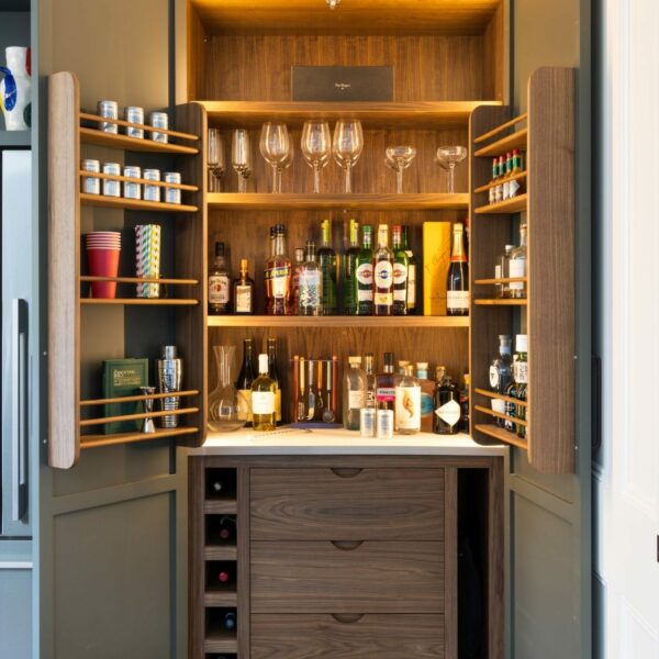 Shaker kitchen larder with doors open, showing bar cabinet, drinks bottles, wine glasses, internal LED lighting. Painted in a dark green-grey colour.