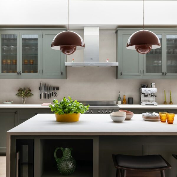 Shaker kitchen island with micro-concrete worktops, a built-in wine cooler, open base shelving, and three walnut bar stools. A range cooker sits against the back wall with a freestanding stainless steel extractor above, surrounded by wall units, some with fluted glass doors.