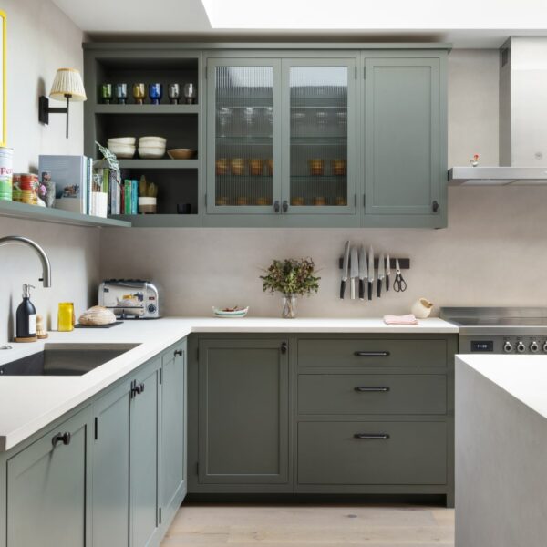 Shaker kitchen island with micro-concrete worktops, a built-in wine cooler, open base shelving, and three walnut bar stools. A range cooker sits against the back wall with a freestanding stainless steel extractor above, surrounded by wall units, some with fluted glass doors.
