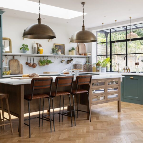 Green Shaker kitchen with a stained oak island featuring glass-fronted drawers and aged brass pull handles. Light quartz worktops extend into the splashback and shelf. Brass accents in the lighting complement the oak herringbone flooring. Large crittal-style windows offer views of the garden.