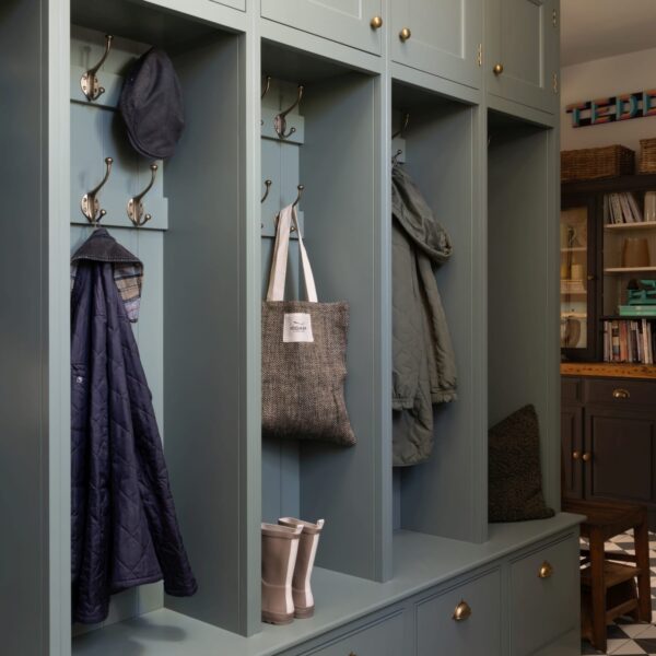 Shaker-style boot room with a bench, drawers and locker-room style cubbies with aged brass hooks. Coats hanging on hooks, wellington boots sit on top of bench. The boot room features a classic black and white checkered floor.