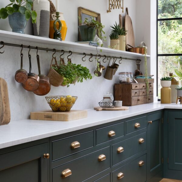 Green shaker kitchen with quartz worktops, splashback, and a matching shelf with a brass hanging rail. Large drawers feature aged brass cup handles. Extending wall lights are mounted above the shelf, which holds ornaments, chopping boards, and fresh herbs.