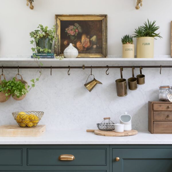 Green shaker kitchen with quartz worktops, splashback, and a matching shelf with a brass hanging rail. Large drawers feature aged brass cup handles. Extending wall lights are mounted above the shelf, which holds ornaments, chopping boards, and fresh herbs.