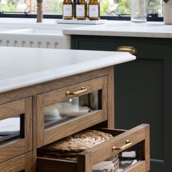 A stained oak island featuring glass-fronted drawers and aged brass pull handles. The light quartz worktops feature an ogee edge.