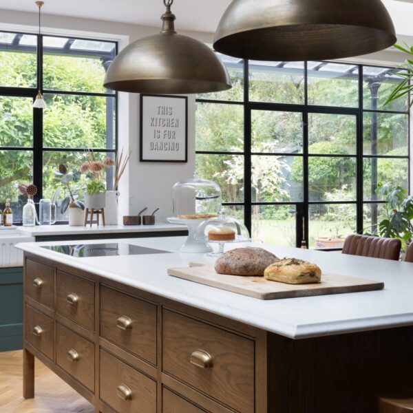 A stained oak kitchen island in a green Shaker kitchen, featuring eight drawers with aged brass pull handles. A vented Bora hob is set into the light quartz worktop with an ogee edge. Oversized brass pendant lights hang above.