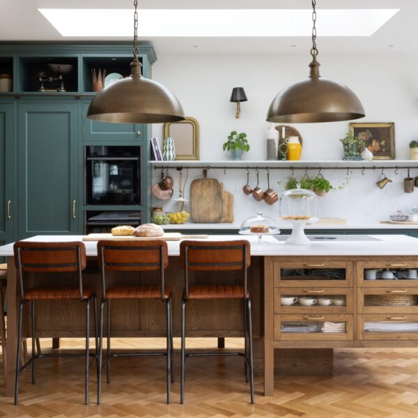 Green Shaker kitchen with a stained oak island featuring glass-fronted drawers and aged brass pull handles. Light quartz worktops extend into the splashback and shelf. Copper pots and fresh herbs hang on S-hooks on a brass hanging rail.