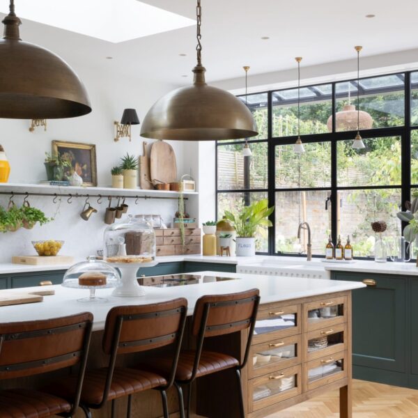 Green Shaker kitchen with a stained oak island featuring glass-fronted drawers and aged brass pull handles. Light quartz worktops extend into the splashback and shelf. Brass accents in the lighting complement the oak herringbone flooring. Large crittal-style windows offer views of the garden.