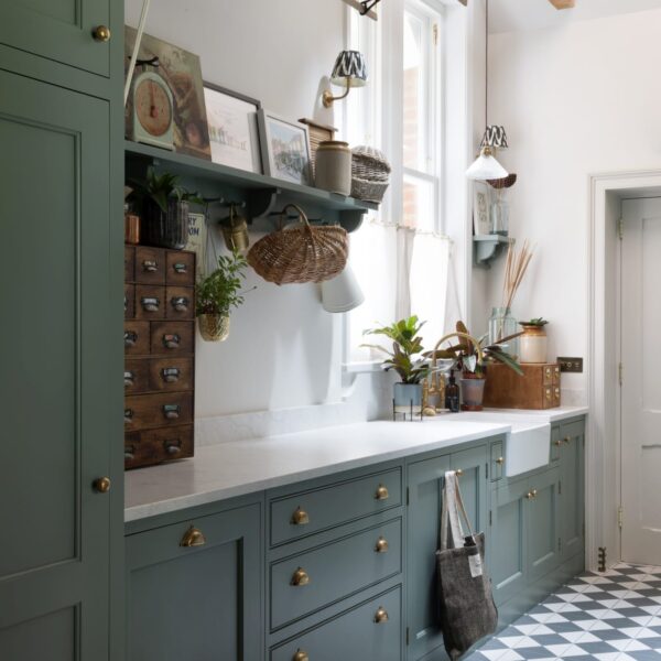 Shaker-style utility room with muted green cabinetry, brass cup handles, and a classic black and white checkered floor. A pendant light hangs from a wooden beam above the sink, positioned in front of a large window with sheer curtains, allowing natural light to brighten the space.