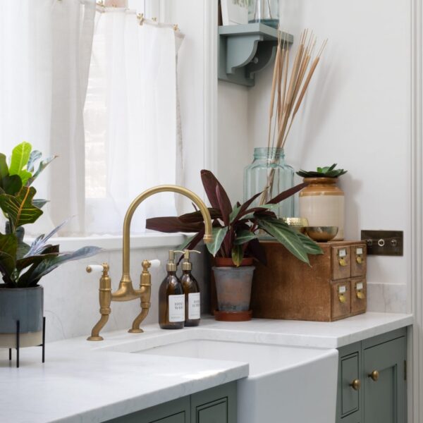 Muted green Shaker utility room with a Belfast sink in front a window with sheer white curtains. An aged brass bridge tap matched the ages brass knobs.