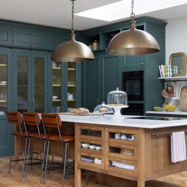 Green Shaker kitchen with a stained oak island featuring glass-fronted drawers and aged brass pull handles. The island has leather bar stools with black metal legs and two wooden stools on the side. A vented hob sits opposite the seating area, with oversized brass pendant lights hanging above. Tall larder cabinets have fluted glass doors.
