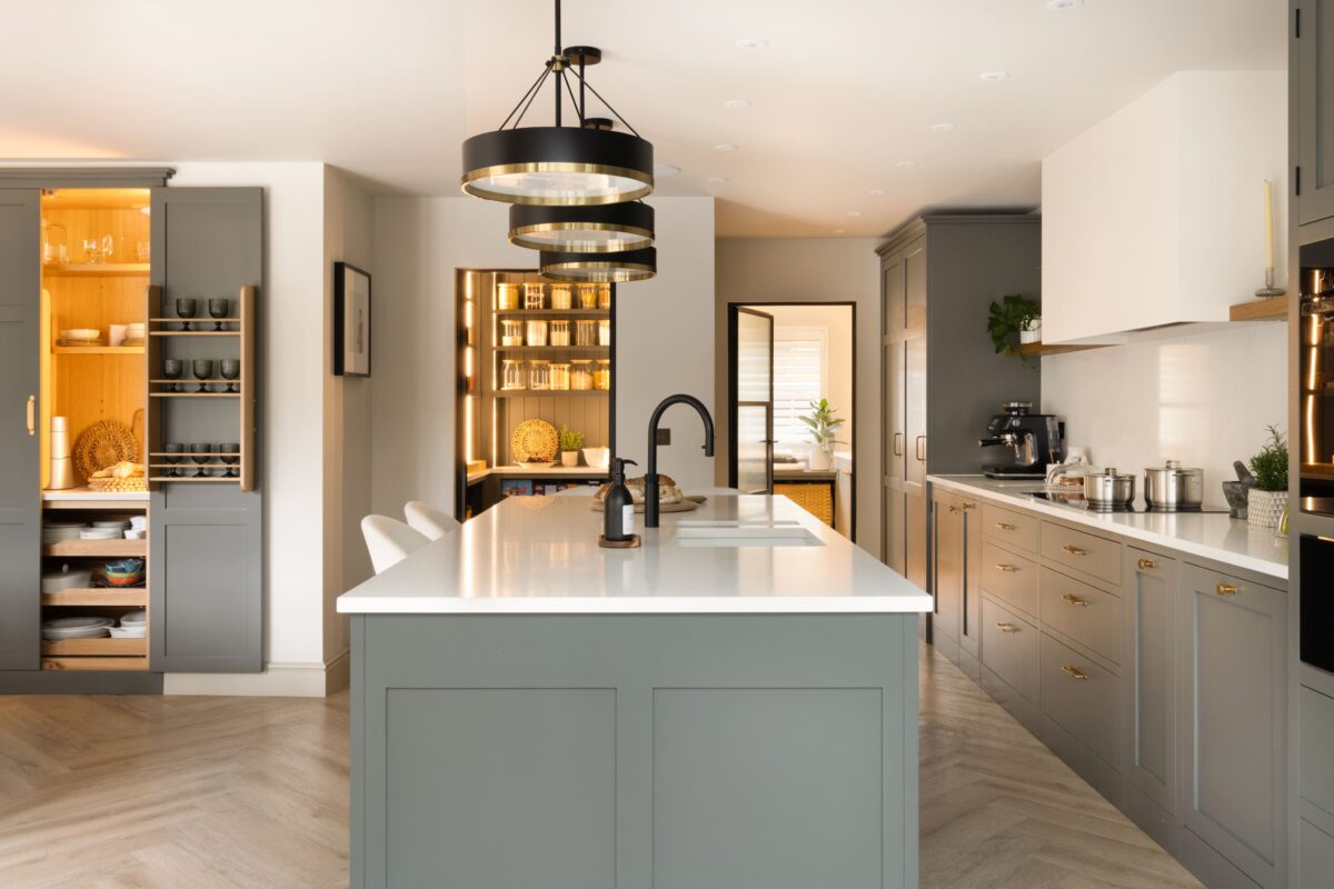 Open-plan kitchen island in sage green with brass detailing, black pendant lighting, and open shelving in a warm oak-lined pantry