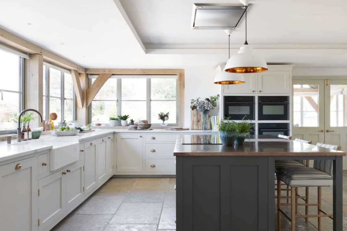 Cream shaker kitchen with dark charcoal island, brass cup handles, Belfast sink, stone flagstone floor, and oak beam ceiling — warm neutral colour palette for a north-facing country kitchen